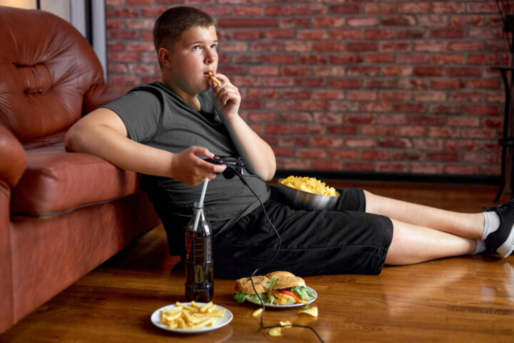 Um menino obeso comendo junk food e jogando videogame em frente à TV